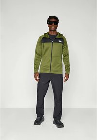 Man staat met zwarte zonnebril, een olijfgroene hoodie met rits en zwarte accenten, zwarte broek en zwarte sneakers tegen een effen achtergrond.
