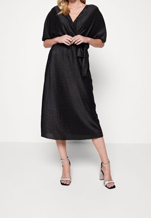 Femme en robe portefeuille midi noire texturée à manches courtes et ceinture, portant des sandales argentées à talons hauts, debout contre un mur blanc.