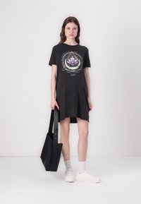 Charcoal gray t-shirt dress with a lotus flower graphic, round neckline, short sleeves, and a curved hem. Model holds a black tote bag.