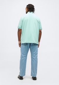 Άνδρας με πλεξούδες, που φοράει κοντομάνικο πόλο σε μέντα πράσινο, ανοιχτό μπλε τζιν και μαύρα παπούτσια, στέκεται με την πλάτη προς τα εμπρός σε απλό φόντο.