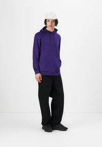 Sudadera púrpura con cordones y bolsillo frontal, combinada con pantalones negros holgados y zapatos negros. El modelo complementó el atuendo con una gorra blanca.