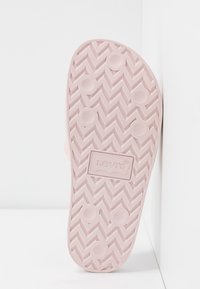 Semelle d’une sandale slide rose Levi’s avec motif de bande de roulement en chevrons et grips circulaires, positionnée verticalement contre un fond blanc d’angle.