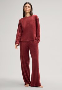 Bordeauxfarbener Loungeset aus weichem, samtigem Stoff. Besitzt ein locker sitzendes Oberteil mit langen Ärmeln und eine weit geschnittene Hose mit elastischem Bund.