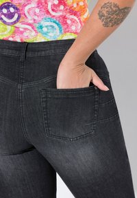Jeans en denim noir avec un design ajusté. Présente une poche arrière avec des coutures visibles et une main placée à l'intérieur. Texture lisse.