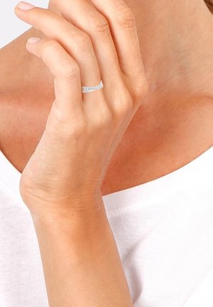 Main à peau claire portant une fine bague en argent à l'annulaire, touchant doucement la poitrine au-dessus d'un haut blanc.