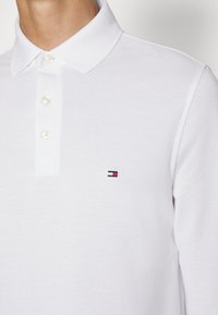 Hvit polo skjorte med teksturert stoff, en knappestolpe og en liten logo i rød, hvit og blå farger på brystet.