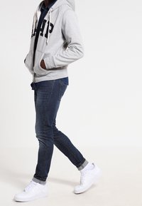 Felpa grigia con zip e lettering "GAP", camicia di jeans blu sotto, jeans scuri e sneakers bianche. La felpa ha coulisse e tasche.