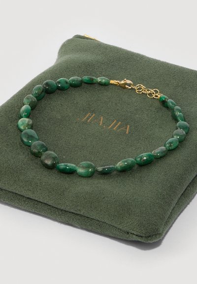 Collana di pietre preziose verdi con perle ovali e tonde, fissata con una chiusura dorata, poggiante su un sacchetto verde scuro.