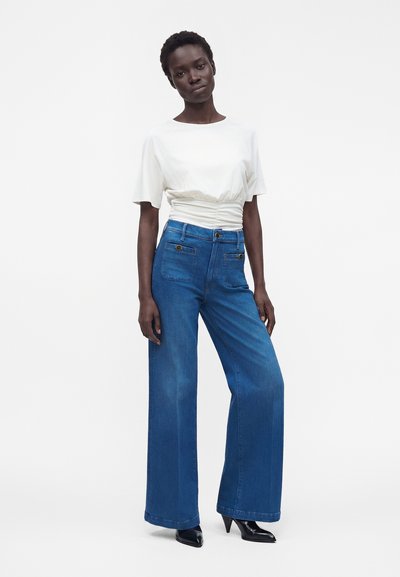 Femme aux cheveux courts portant un haut blanc froncé à manches courtes, un jean bleu taille haute à jambes larges et des talons noirs pointus, debout devant un fond uni.