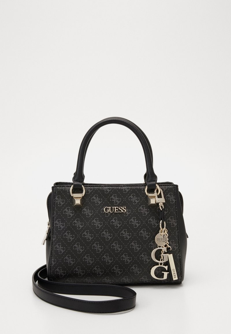 Sac à main noir Guess avec double poignée, motif monogramme, bandoulière amovible et breloques logo dorées sur fond blanc.