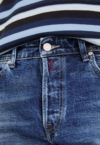 Jeans de mezclilla azul oscuro con un diseño ajustado, que cuentan con un botón plateado, costuras rojas y bolsillos visibles. Una camiseta a rayas asoma por encima.