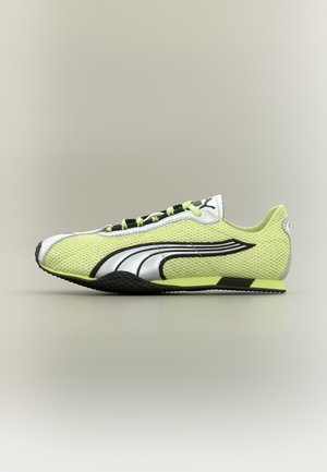 Zapatilla deportiva de malla verde claro y blanca con suela negra y blanca, cordones negros y un logo audaz en negro y plata en el lateral.