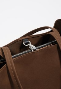 Ruda audinio pirkinių maišelis su sidabriniu užtrauktuku, juodais odiniais akcentais ir dvigubai siūtais rankenomis, demonstruojantis grakščią, minimalistinę dizaino estetiką.