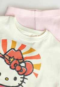 Kremowa koszulka z grafiką Hello Kitty, która ma czerwoną czapkę i wstążkę, warstwowa na jasnoróżowych, prążkowanych spodniach.