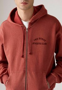 Felpa rossa con zip, realizzata in tessuto morbido, dotata di tasca frontale, cordini e logo "TWO HORSE ATHLETIC CLUB" in stampa scura.