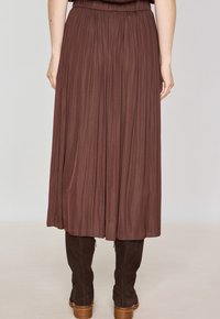 Gonna plissettata marrone con elastico in vita, tessuto fluido, lunghezza ginocchio, indossata con stivali scamosciati scuri fino al ginocchio.