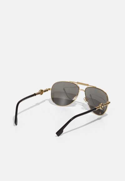 Kultaiset metalliset aviator-aurinkolasit harmailla linsseillä, joissa on mustat asetattihaarat ja monimutkaiset metalliosat nenäosassa ja sivuilla.