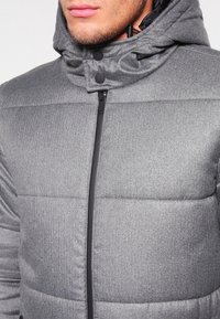 Grå polstret jakke med lynlåslukning og hætte, der har et struktureret stof, sort lynlås og trykknapper.