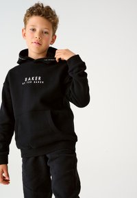 Must kapuutsiga pusa, millel on valge tekst "BAKER BY TED BAKER". Omaduseks on esitasku ja soonikkoes varrukasuu, kantud sobivate mustade pükstega.