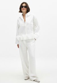 Weißes Button-up-Hemd mit Rüschensaum, kombiniert mit weißen Leinenhosen und weißen durchlöcherten Slippern. Modell trägt dunkle Sonnenbrillen.