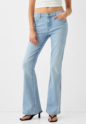 Femme portant un jean flare bleu clair, un débardeur court blanc et des sandales noires à talons, debout avec une main dans la poche.