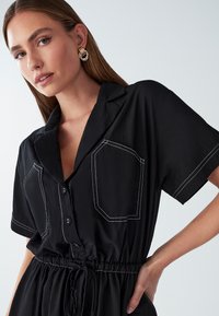 Donna con lunghi capelli castani che indossa orecchini a cerchio dorati e un vestito nero con bottoni, caratterizzato da cuciture bianche e una vita sottolineata, su uno sfondo semplice.