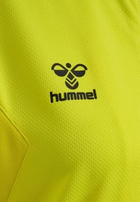 Gult sportsjersey med en tekstureret, åndbar stof. Logoet "hummel" og et stiliseret design er trykt i sort.