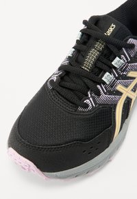 Černá sportovní bota s prodyšným svrchním materiálem, zlatými a bílými detaily, texturovaným designem a světle růžovou gumovou podrážkou. Obsahuje šněrování.