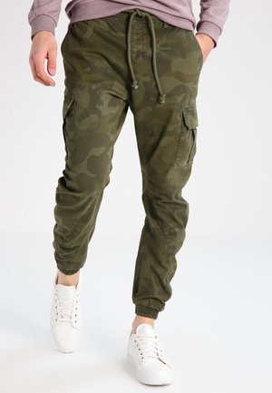 Hombre con pantalones jogger cargo de camuflaje verde oliva y zapatillas blancas, caminando con las manos parcialmente en los bolsillos.