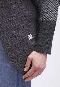 Handen vilar mot höften, klädd i en mörk, texturerad stickad tröja med G-Star Originals-märke och blå denimjeans.