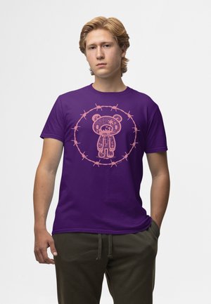 Giovane uomo con capelli biondi che indossa una maglietta viola con una grafica di un orsetto rosa circondato da filo spinato, in piedi con una mano in tasca.