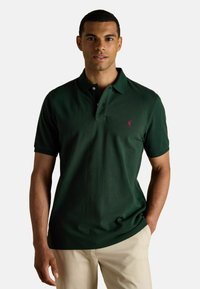 Tume roheline polo särk, mis on valmistatud puuvillast, kolme nööbiga kaelusega, lühikeste varrukatega ja vasakul rinnal väikese roosa logoga.