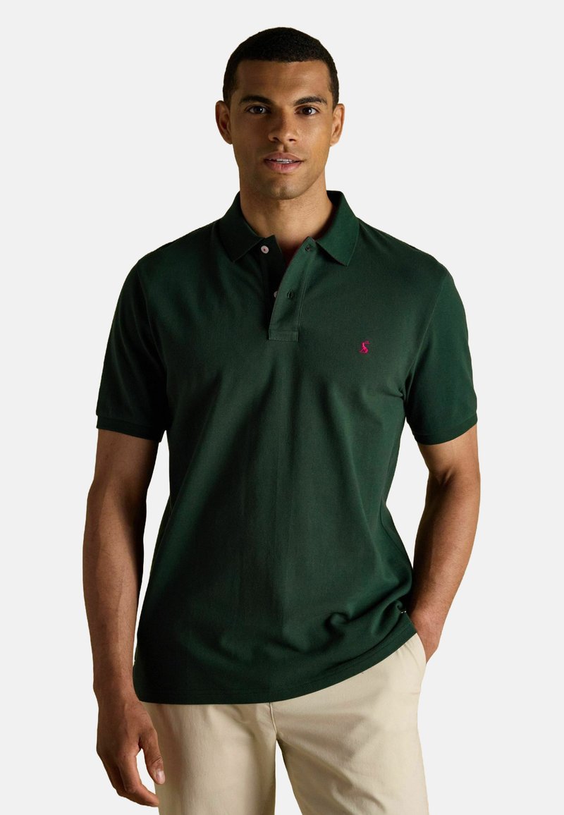 Tume roheline polo särk, mis on valmistatud puuvillast, kolme nööbiga kaelusega, lühikeste varrukatega ja vasakul rinnal väikese roosa logoga.