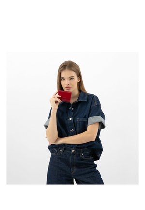 Denim-Hemd mit hochgeschlagenen Ärmeln, kombiniert mit dunkelblauen Jeans. Hält eine kleine rote Geldbörse, vor einem schlichten weißen Hintergrund.