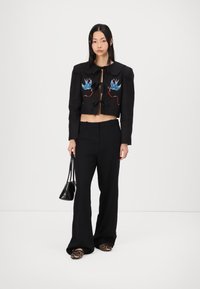 Giacca nera cropped con cigni blu ricamati, legata con fiocchi. Abbinata a pantaloni neri a gamba larga e scarpe slip-on con stampa leopardata.