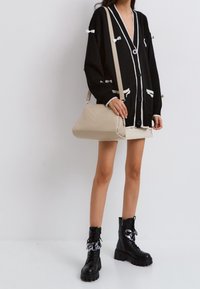 Cardigan noir avec accents blancs et détails de nœud ; sac bandoulière beige à motif chevron ; bottes noires à lacets avec semelles épaisses.