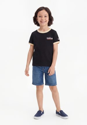 Bambino sorridente in piedi indossa una t-shirt nera Champion con finiture beige, pantaloncini di jeans e scarpe slip-on blu navy su sfondo bianco.