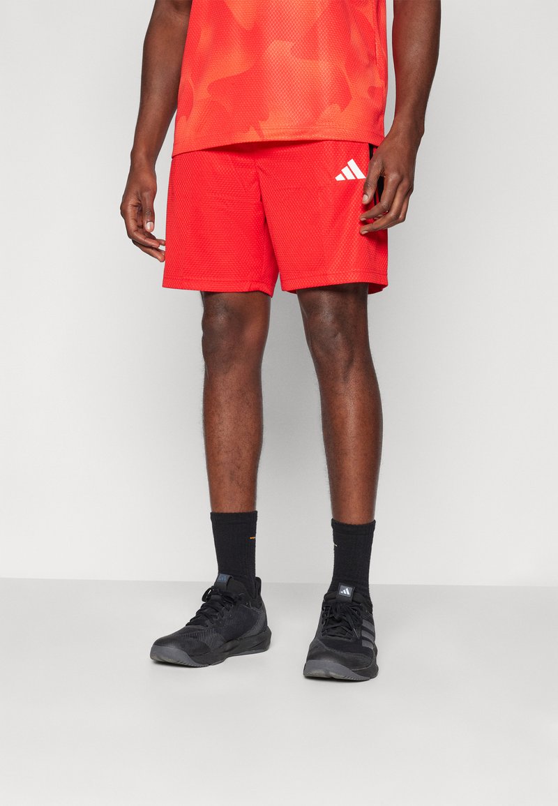 Rdeče atletske kratke hlače z mrežasto teksturo, z belim logotipom Adidas na strani. V paru z črnimi čevlji in nogavicami.