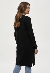 Crna pletiva cardigan sa teksturiranim uzorkom, dugim rukavima i bočnim prorezima. Nositeljica ga nosi preko svijetloplavih traperica.