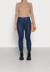 Hnědý žebrovaný rolák, tmavě modré džíny s vysokým pasem a černé kotníkové boty. Béžová crossbody kabelka s řetízkovým popruhem.