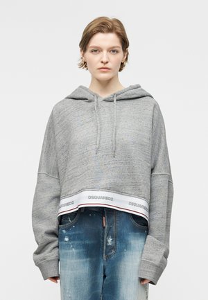 Γυναίκα με κοντά μαλλιά που φοράει μεγάλα κρίκους σκουλαρίκια, γκρι κοντό φούτερ με τη σήμανση DSQUARED2 και ταλαιπωρημένο μπλε τζιν.