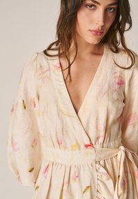 Jonge vrouw met golvend bruin haar, gekleed in een lichtbeige bloemenwikkeljurk met pofmouwen en een gestrikte tailleband.