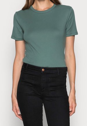 Vihreä puuvillainen t-paita, jossa on lyhyet hihat ja pyöreä pääntie, yhdistetty korkeavyötäröisiin mustiin farkkuihin, joissa on etutaskut ja nappi.