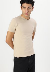 Camiseta de algodón beige con mangas cortas, escote redondo, costuras mínimas y un pequeño logo triangular rojo en el pecho. Combinada con jeans oscuros.