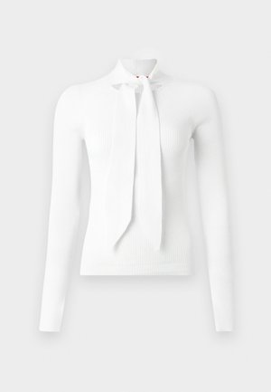 Hvit rysjede langermet topp med høy hals og foran knyte-detalj. Har en nøkkelhullskjæring ved halsen og en myk tekstur.