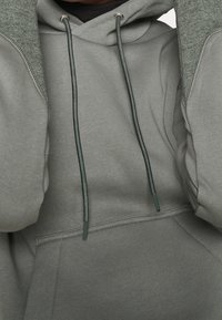 Felpa grigia con cappuccio, caratterizzata da una sovrapposizione in maglia verde testurizzata sulle spalle, stringhe regolabili e una tasca frontale.