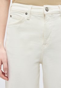 Gros plan sur une personne portant un jean denim blanc taille haute avec une poche avant visible et un bouton métallique estampillé Lee.