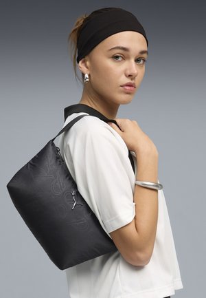 Joven con diadema negra y joyas plateadas lleva bolso de hombro negro con patrón sutil sobre fondo gris.