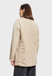 Béžová péřová bunda se rovný designem, vyrobená z hladkého, lesklého materiálu a s mírně polstrovanou texturou. Mezi detaily patří boční kapsy.