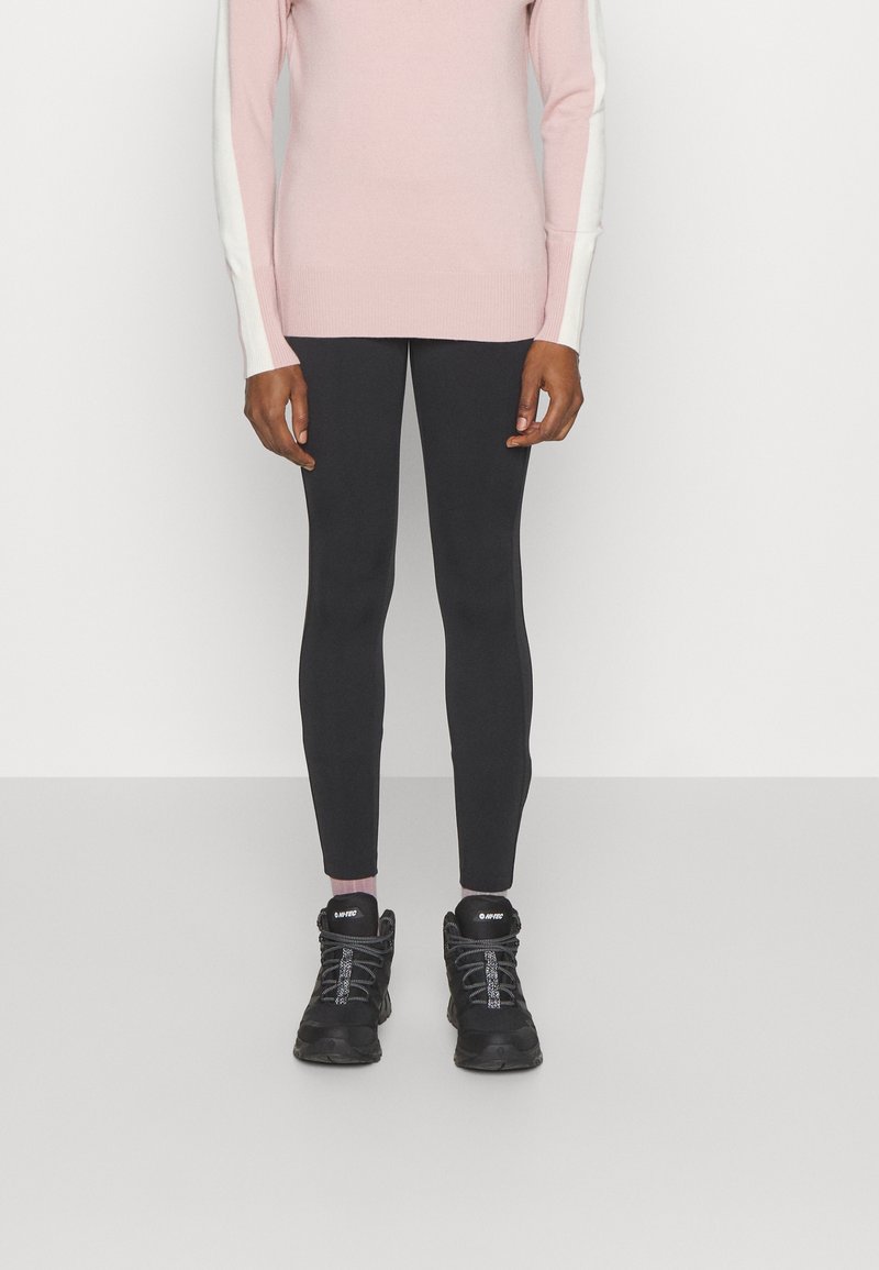 Une personne se tient debout, portant un pull rose avec des manches blanches, des leggings noirs et des chaussures de sport noires, sur un fond gris clair.